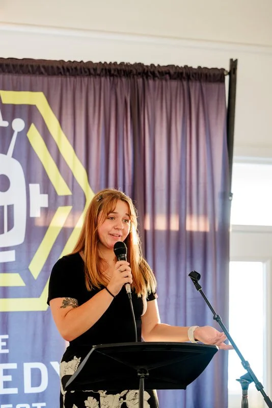 A young woman with red hair speaking into a microphone at a podium, with a purple curtain and a large purple and yellow logo in the background, in a well-lit indoor setting.