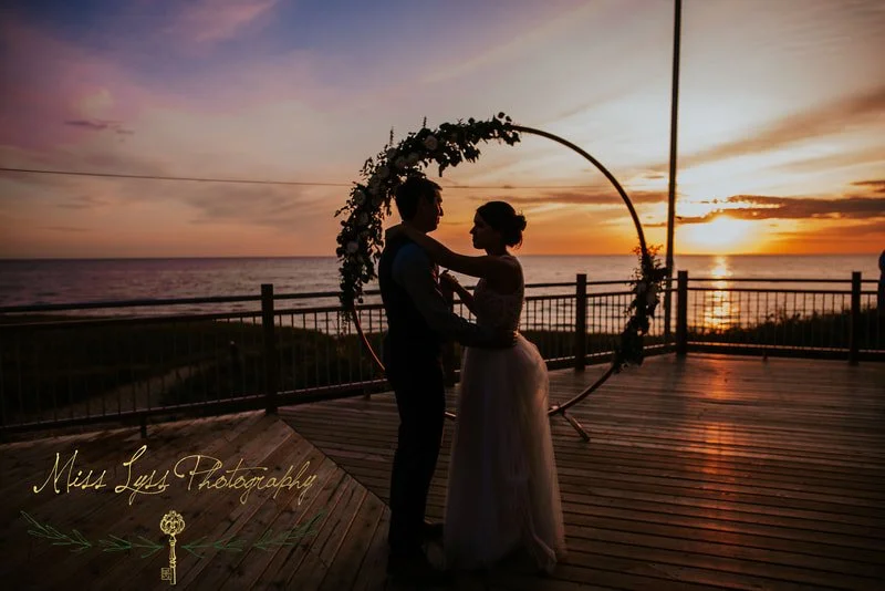 A couple dancing outdoors at sunset with an ocean view in the background, framed by a circular floral arch on a wooden deck.