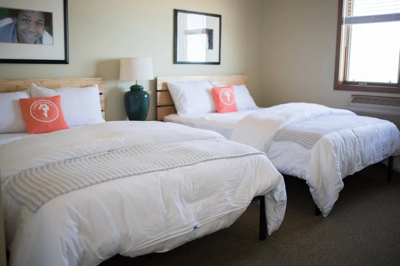 A bedroom with two beds, each with white bedding, and a matching orange pillow featuring a white logo, a nightstand with a green lamp between the beds, and artwork on the wall, a window on the right allowing natural light.