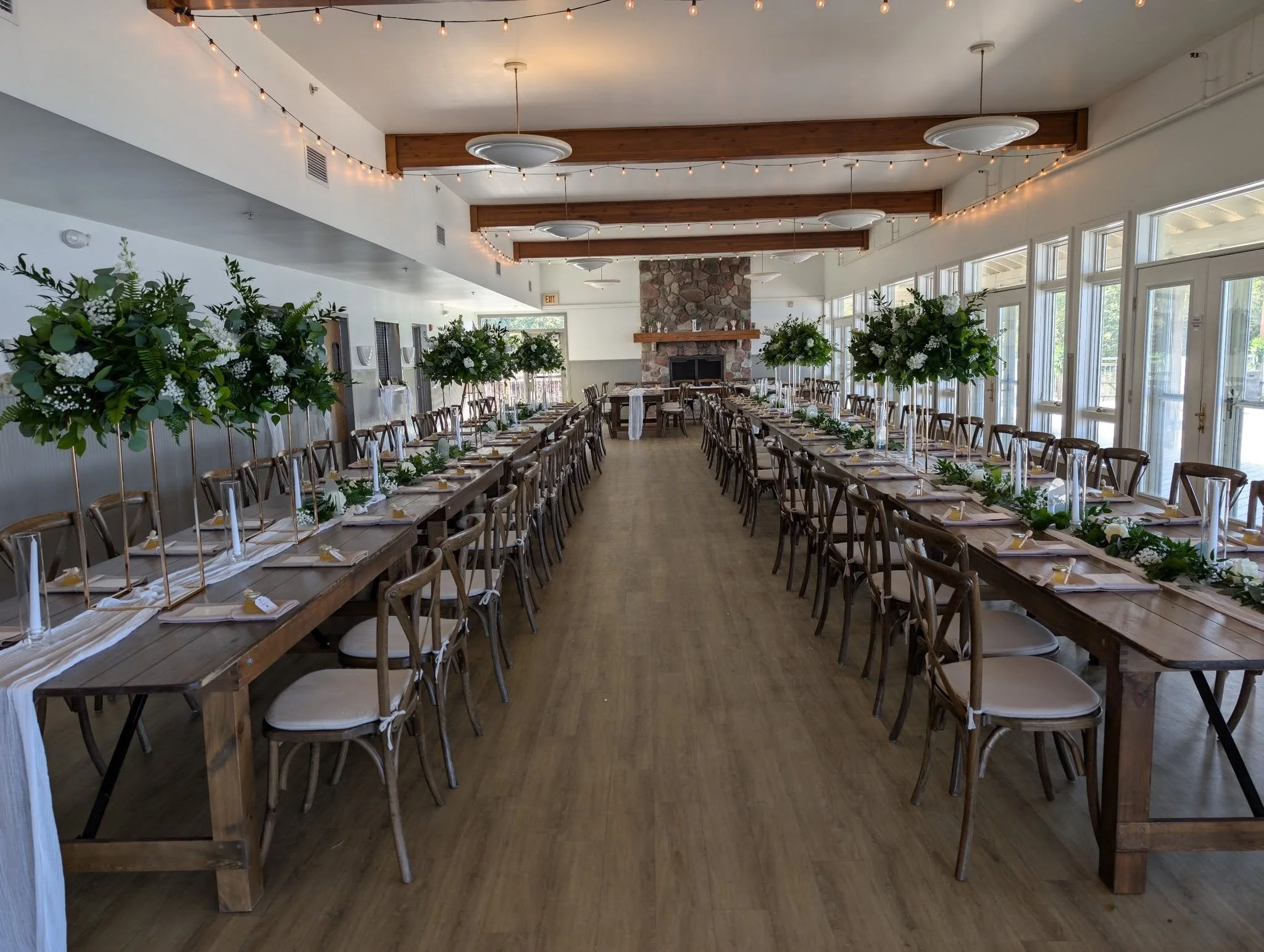 Two rows of farm tables with matching chairs set up in the Loeks Retreat Center at Camp Blodgett for a special event rental. There is a head table set in front of a fireplace..