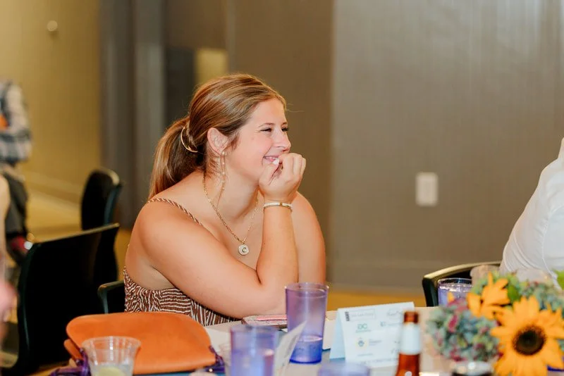 A young woman with brown hair in a ponytail, smiling and laughing with her hand near her mouth at a table decorated with flowers and drinks in a banquet hall.