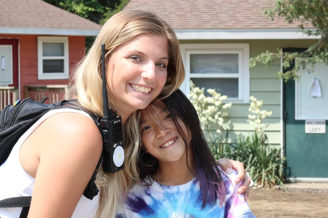 Two young women smiling and hugging outdoors in front of a house with garden plants.