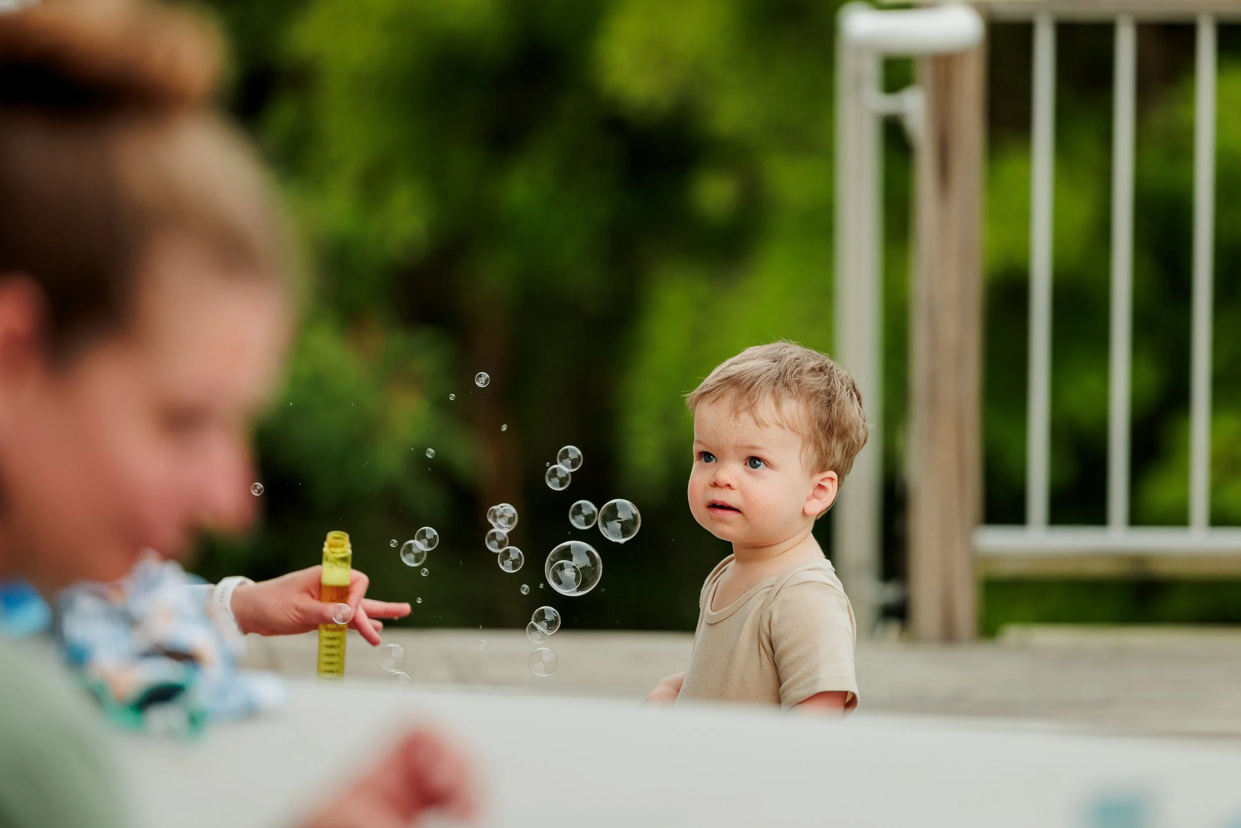A young boy with short blonde hair and blue eyes looking at a woman blowing soap bubbles outdoors on a wooden deck with green trees in the background.
