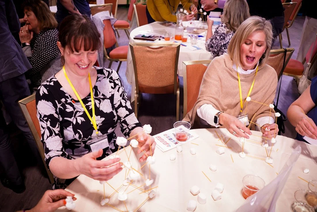 Two women seated at a table, engaging in a marshmallow and spaghetti tower building activity during a group event or workshop, with about a dozen people in the background.