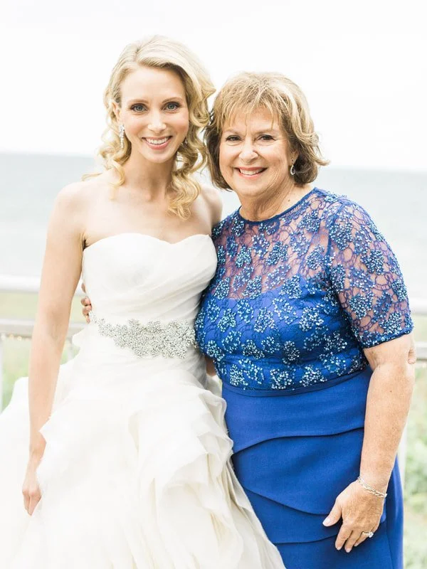 Two women, a bride in a white wedding dress and an older woman in a blue dress, smiling together outdoors near the beach.