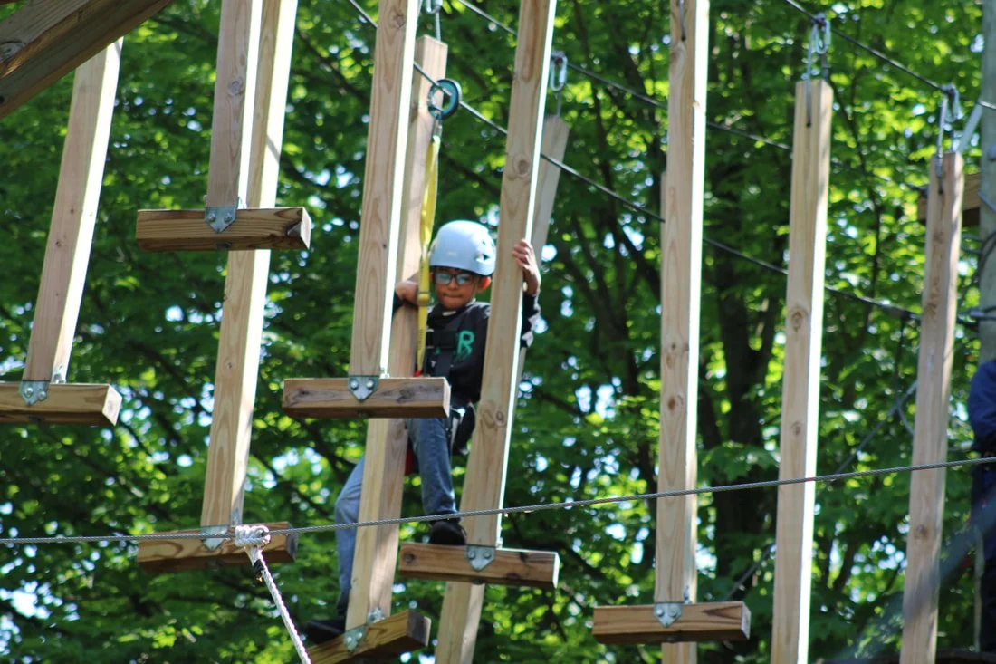 Child wearing a helmet and harness crossing a wooden rope bridge at a high ropes course in a forest.