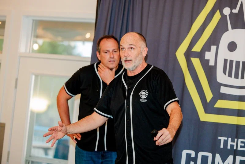 Two men wearing black shirts with white trim, one speaking and the other listening, on a stage with a backdrop featuring a stylized robot and the word 'COMEDY'.