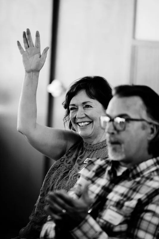 A woman smiling and raising her hand in a meeting or class, sitting next to a man with glasses looking at his phone in an indoor setting.