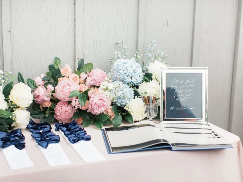 A table decorated with pink, white, and blue flowers, a framed sign, an open guest book, and several navy blue ribbon awards with white tags.