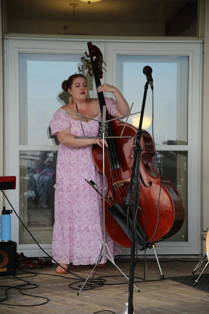 A woman in a pink floral dress playing a double bass on a stage with microphones and musical equipment.