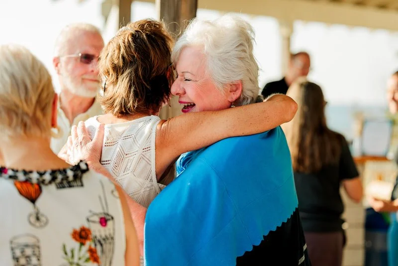 Two elderly women hugging and smiling at an outdoor gathering, with other people in the background.