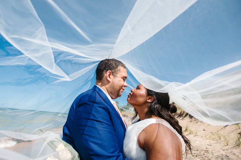 A couple in wedding attire standing under a white fabric canopy on a beach, facing each other with smiles.