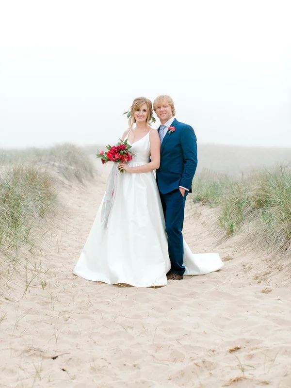 Bride and groom standing on sandy beach with grass on sides, photographed on their wedding day.