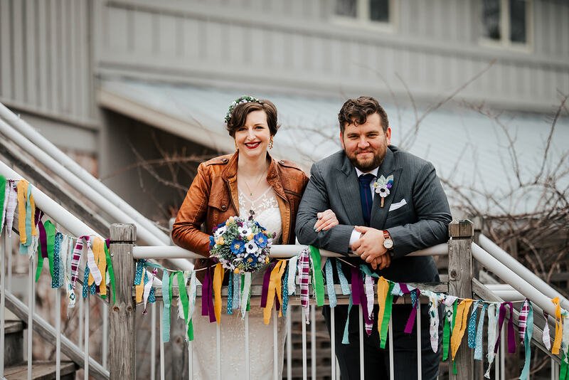 A smiling bride and groom leaning on a bridge railing decorated with colorful hanging ribbons, outdoors with a building in the background during a wedding celebration.