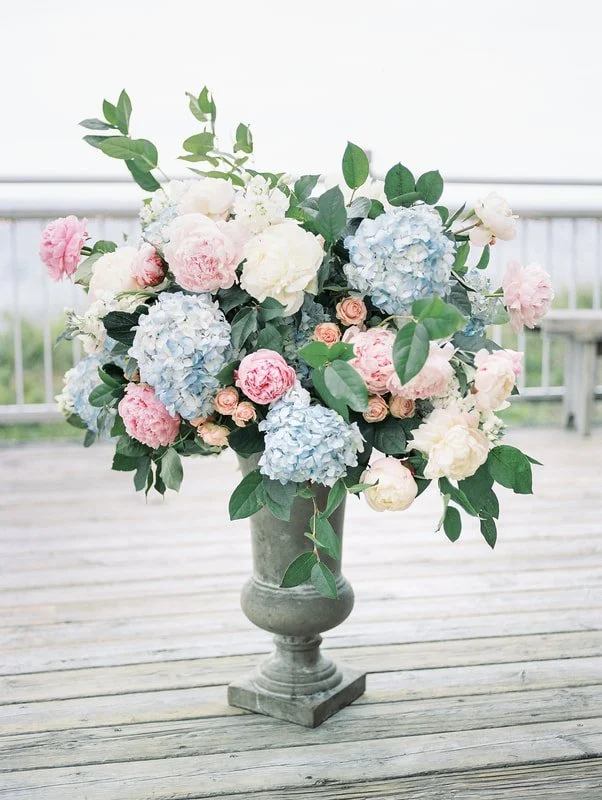 A large bouquet of pink, white, and light blue hydrangeas with green foliage in a gray vase on a weathered wooden table outdoors.