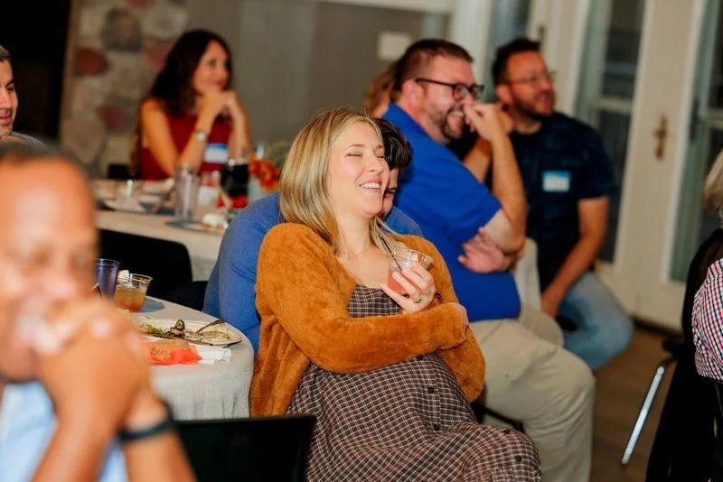 People sitting and laughing at a social gathering or party, with a woman in the foreground holding a drink and smiling.