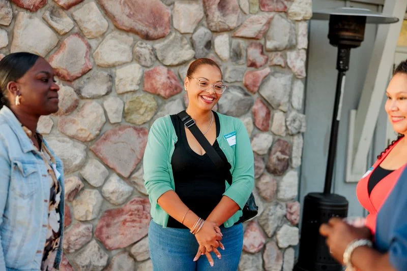 Three women talking and smiling outdoors, with a stone wall in the background.
