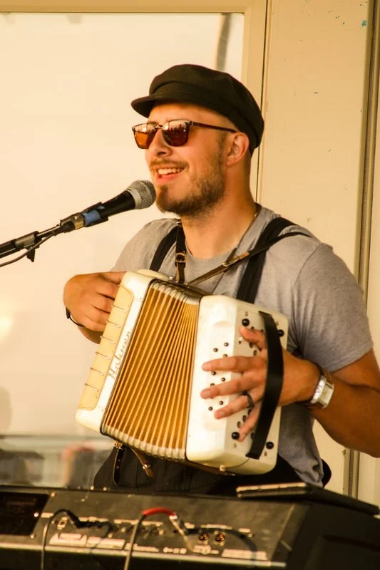 A man wearing sunglasses and a black hat playing an accordion while singing into a microphone.