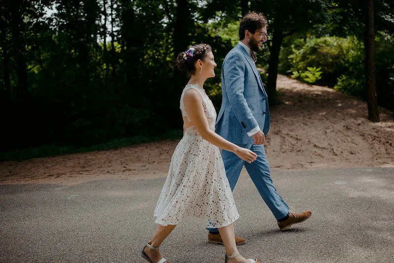 A man and a woman walking together outdoors on a paved path, surrounded by trees and greenery. The woman is wearing a white lace dress and the man is in a light blue suit.