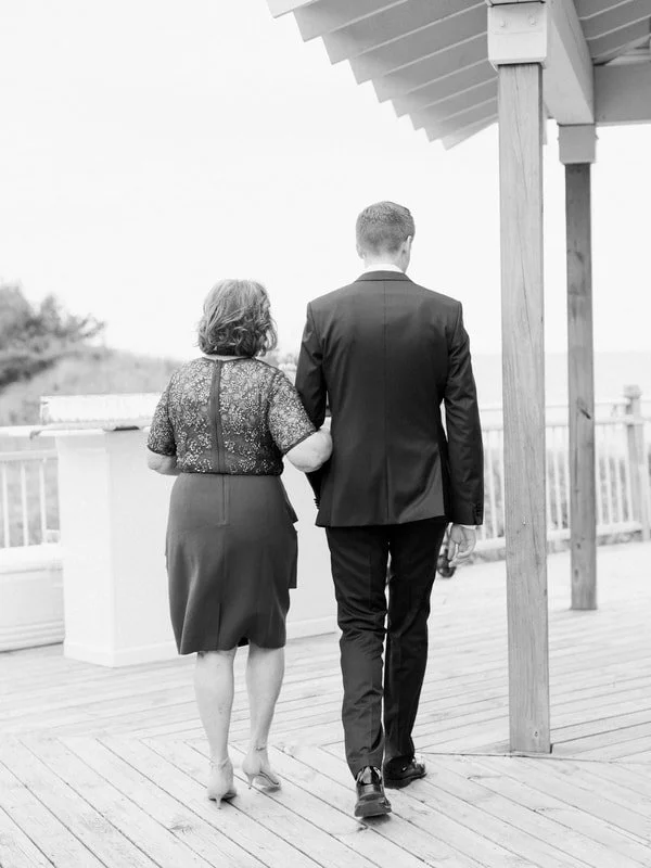 A young man in a suit walking arm-in-arm with an older woman in a dress on a wooden deck, seen from behind.