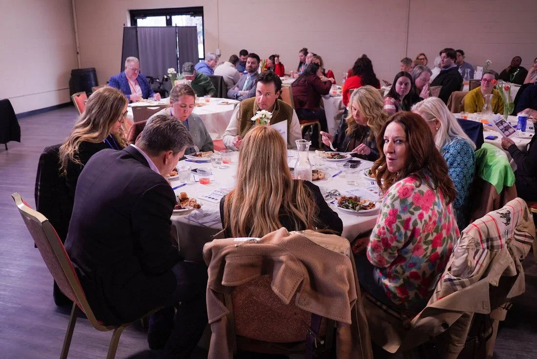 People sitting around a circular dinner table at an indoor event or conference, eating and talking.