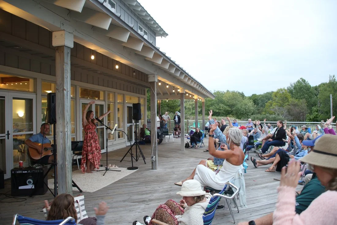 Outdoor concert on a wooden deck with a female singer and a guitarist, audience members seated in chairs raising their hands, in a natural setting with trees in the background at dusk.