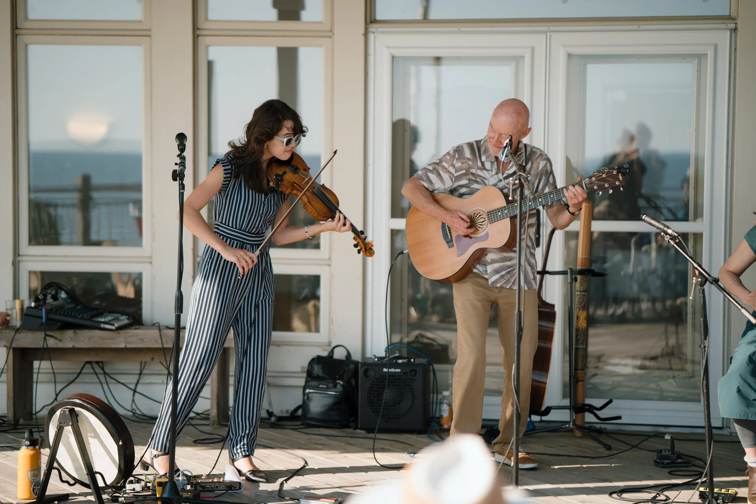 A man and woman performing music outdoors on a wooden deck, with the woman playing a violin and the man playing an acoustic guitar, with microphone stands and musical equipment around them.