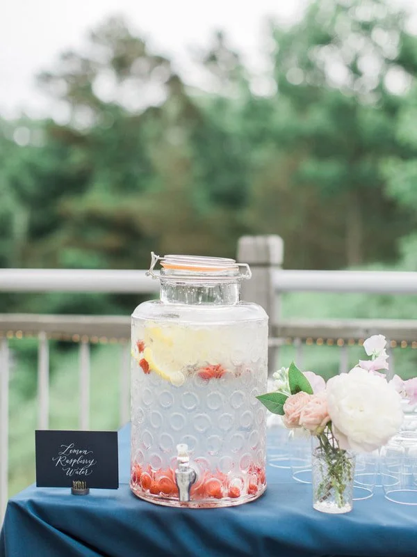 A large glass jar of lemon raspberry water on a table with a spigot, next to a small sign and a flower arrangement.