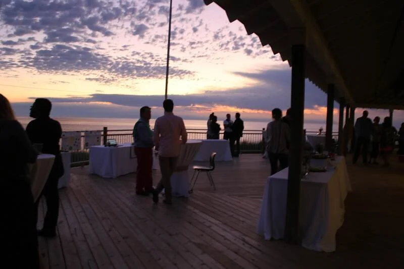 People gather on a wooden deck overlooking the ocean at sunset, some engaged in conversations and others enjoying the view, with tables covered in white cloths set up for an event.