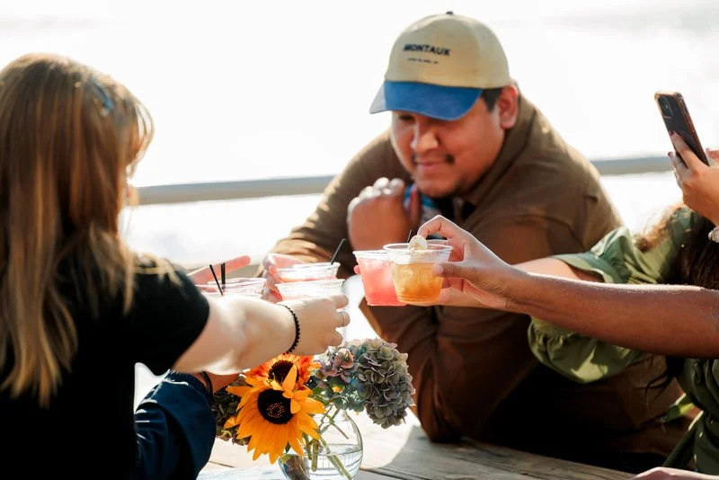 People gathered around a table outdoors, sharing drinks, with flowers in a vase and a scenic water view in the background.