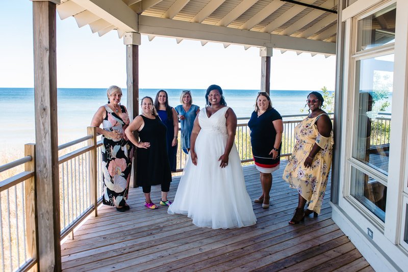 Group of seven women standing on a wooden balcony by the beach, one woman in a white dress, others in colorful dresses, smiling for a photo