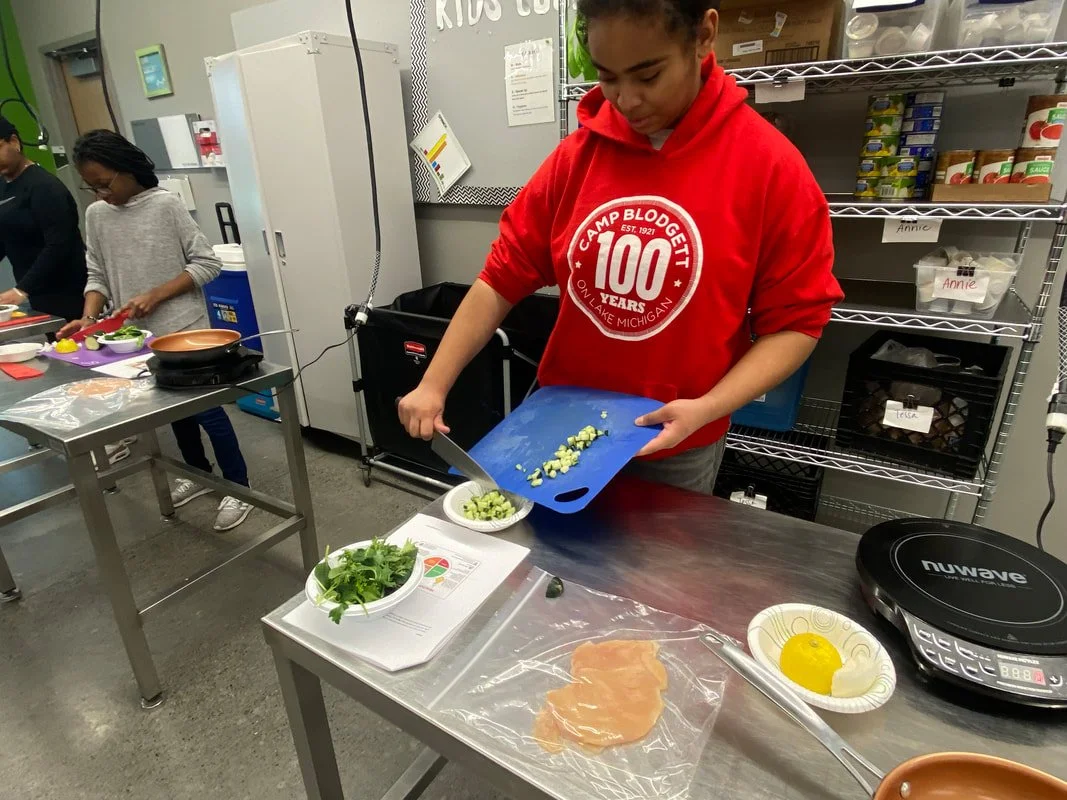 A girl wearing a red Camp Blodgett sweatshirt chopping green onions on a blue cutting board, with a plate of greens and an egg on the counter in front of her. Other people are cooking in the background.