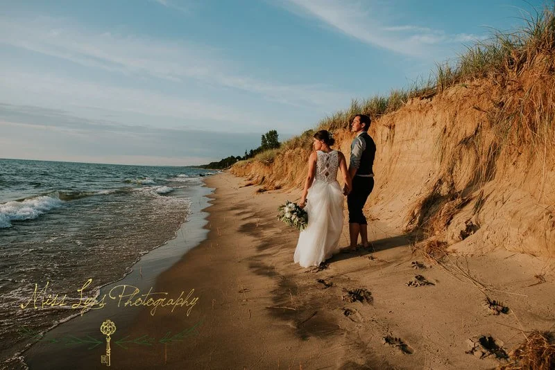 A bride and groom standing on a sandy beach, holding hands, with the bride holding a bouquet of flowers, as they face the ocean under a partly cloudy sky during sunset.