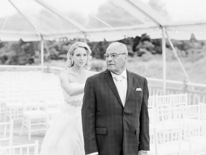 A bride in a wedding dress stands behind an older man in a suit under a tent at an outdoor wedding ceremony.