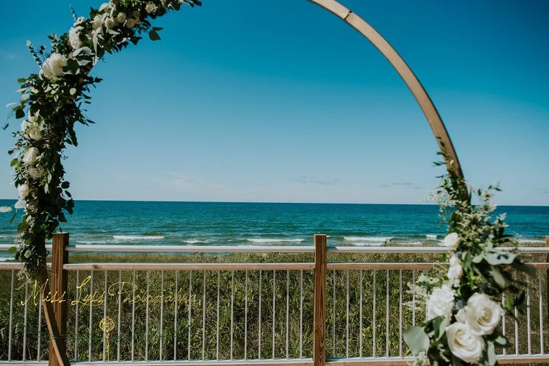 Wedding arch decorated with white flowers and greenery overlooking the ocean, set on a wooden deck.