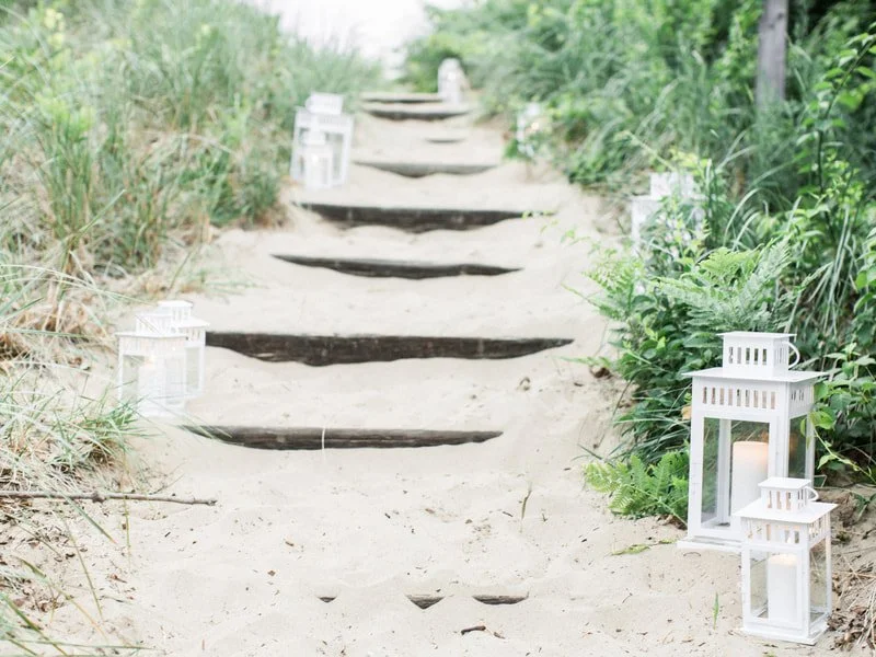 Sandy pathway with wooden steps, white lanterns, and green foliage on both sides, leading to a bright background.