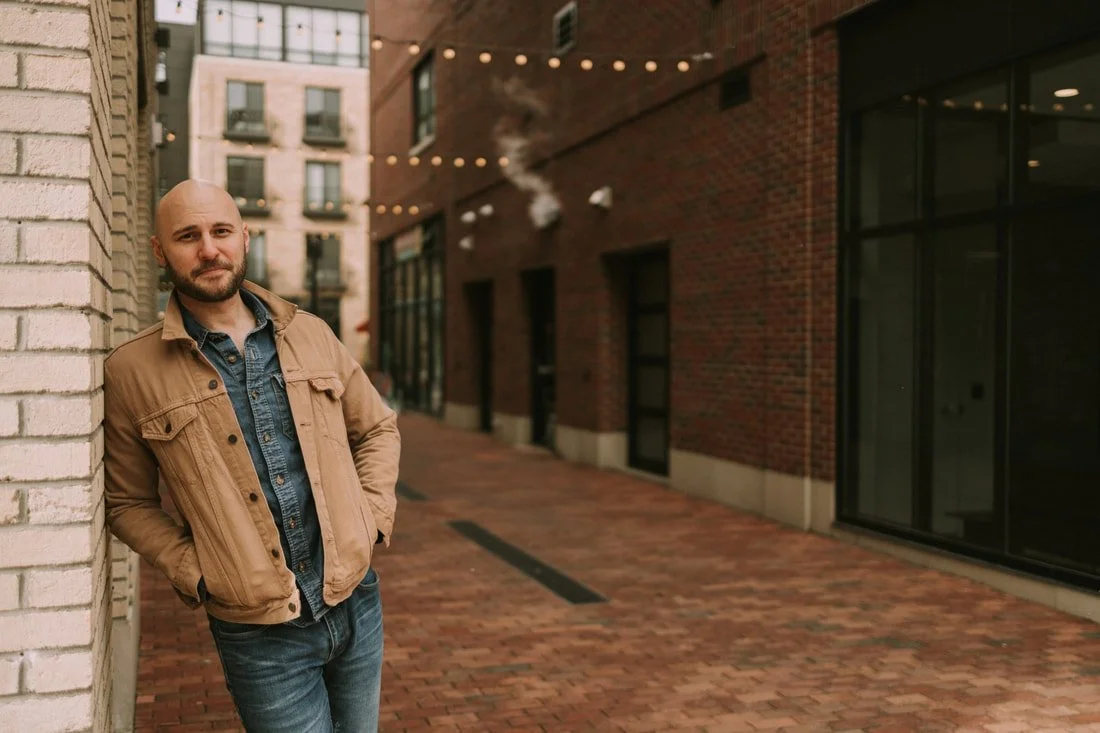 A man with a beard leaning against a brick wall in an urban alleyway, wearing a tan jacket and blue jeans, with outdoor string lights overhead and a building with windows behind him.