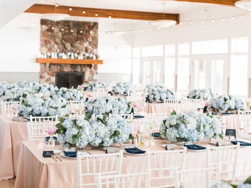 Reception hall decorated for a wedding with round tables covered in blush pink tablecloths, each adorned with large flower arrangements of white hydrangeas and greenery, set with navy blue napkins, glassware, and small chalkboard signs. The room feat