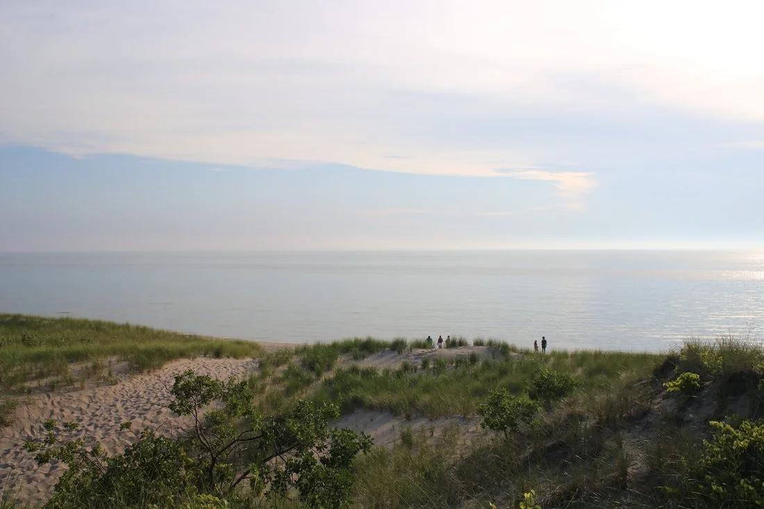 Beach scene with sand dunes, green plants, and a calm ocean under a partly cloudy sky, with a few people near the water.