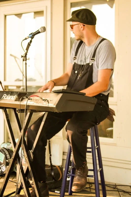 A man wearing glasses, a black hat, gray t-shirt, and overalls playing a keyboard on a stool, with a microphone on a stand nearby.