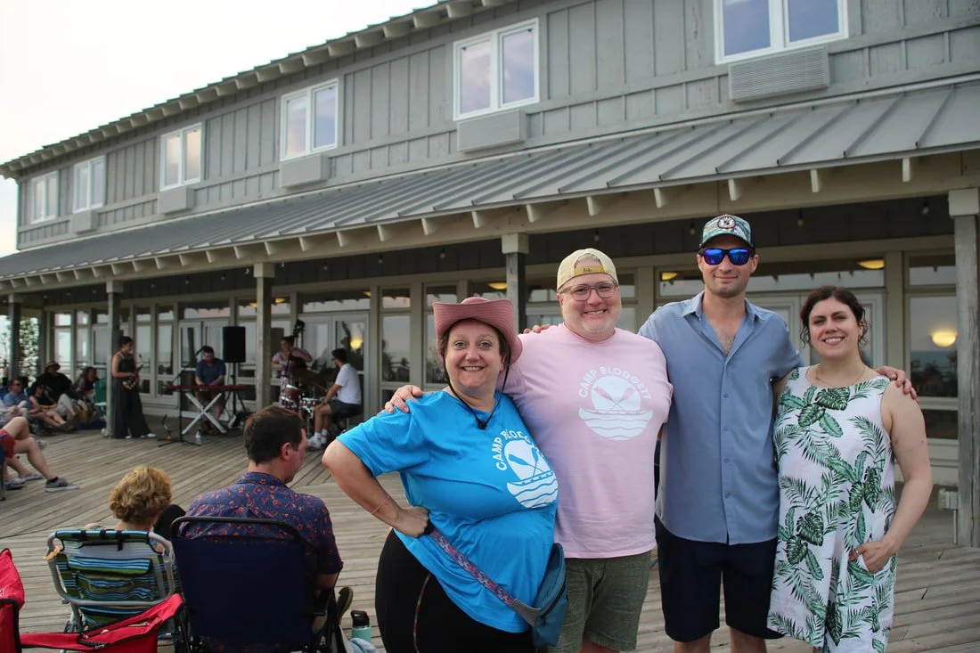 Four people standing on a wooden deck outside a large building, posing for a photo and smiling. Behind them, other people are seated and standing, with some musicians playing in the background.