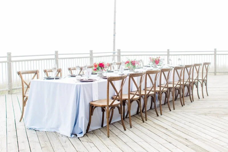 Long dining table with a white tablecloth, floral centerpieces, and place settings, set on an outdoor deck with a white railing.