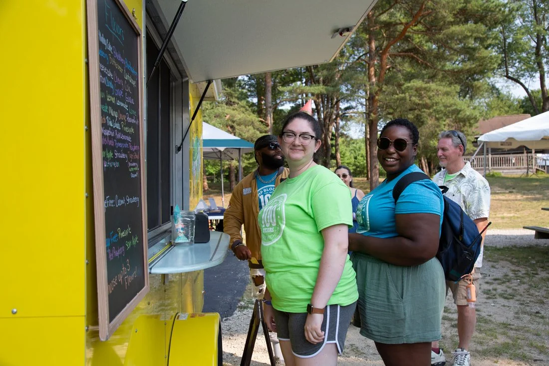 People standing in line at a food truck outdoors on a sunny day, with trees and tents in the background.