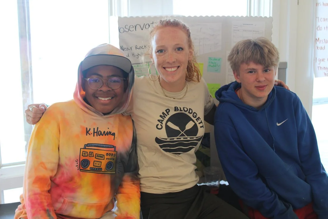 Three smiling teenagers sitting close together indoors, with a large poster behind them displaying handwritten notes and text. The girl in the middle has red hair and is wearing a Camp Blodgett t-shirt, the boy on the right has blond hair and is wear