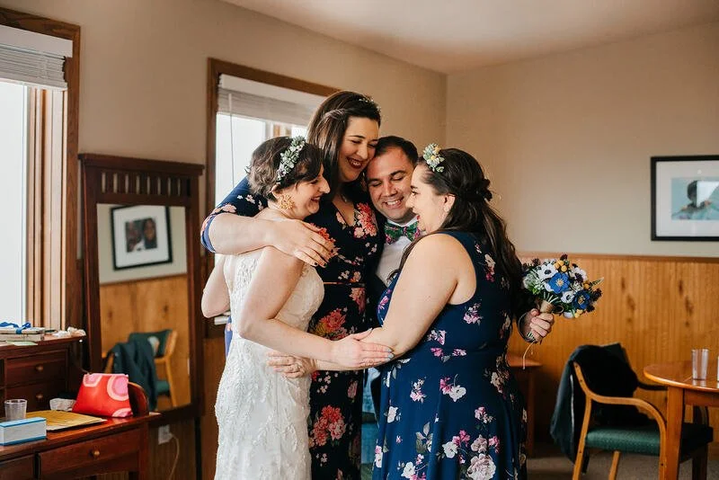 Four people, including a bride in a white lace wedding dress, hugging and smiling, inside a cozy room during a wedding celebration. They are dressed in floral attire, with one holding a bouquet of flowers.
