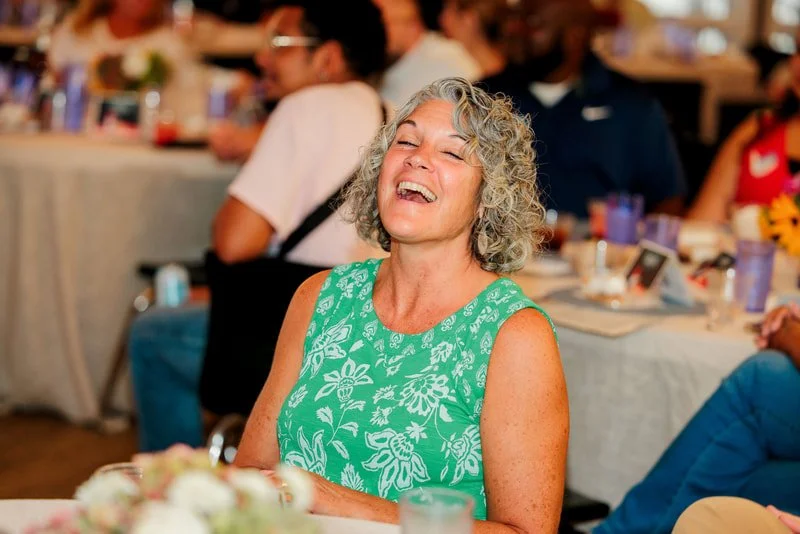 A woman with curly gray hair wearing a green sleeveless dress with white floral pattern is smiling and laughing at a social gathering.