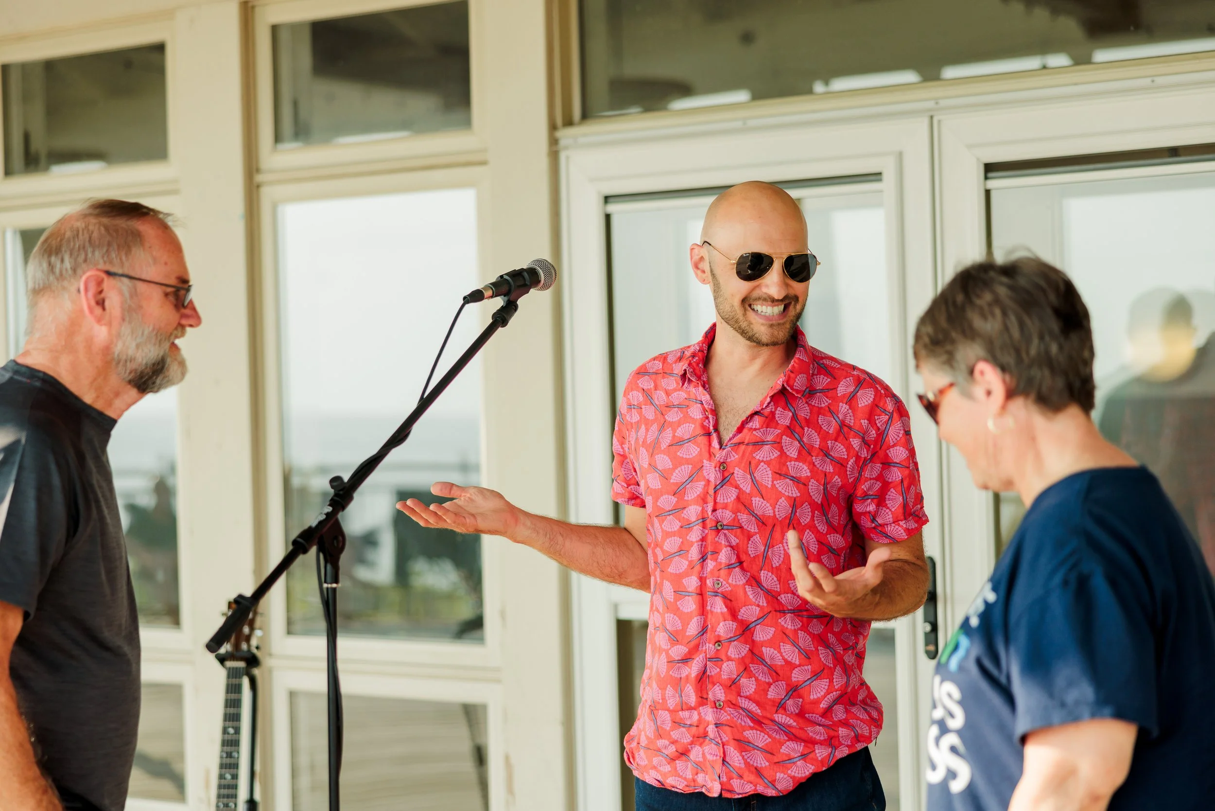 Three people standing together near a microphone, engaged in conversation and smiling. The person in the middle is a bald man wearing sunglasses and a red floral shirt. The person on the left has a beard, glasses, and wears a black shirt. The person 