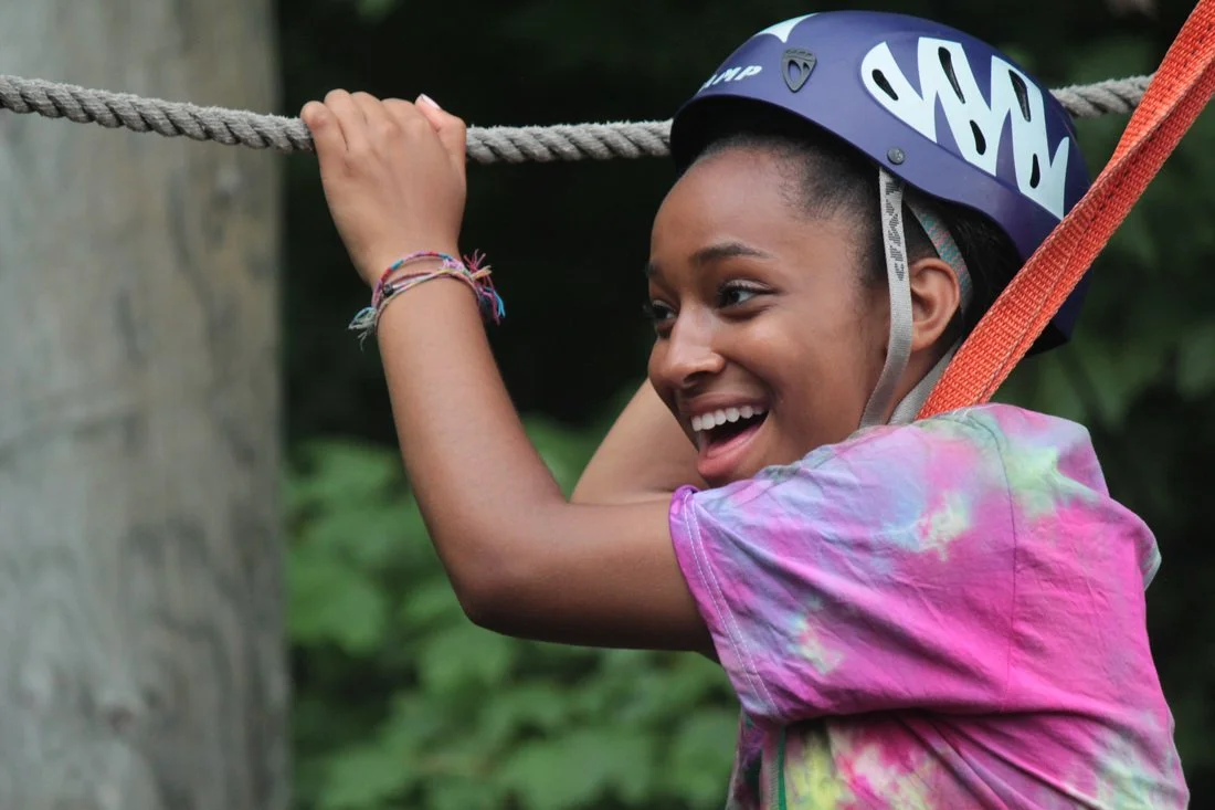 Young girl wearing a bike helmet, hanging on a rope in an outdoor adventure setting, smiling and enjoying herself.