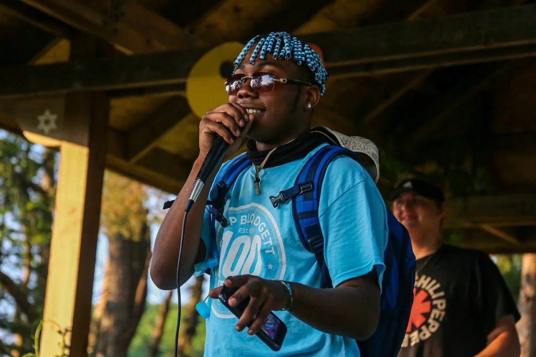 A person with blue braids, sunglasses, and a blue shirt holding a microphone, standing outside with a backpack on, smiling at an outdoor event.