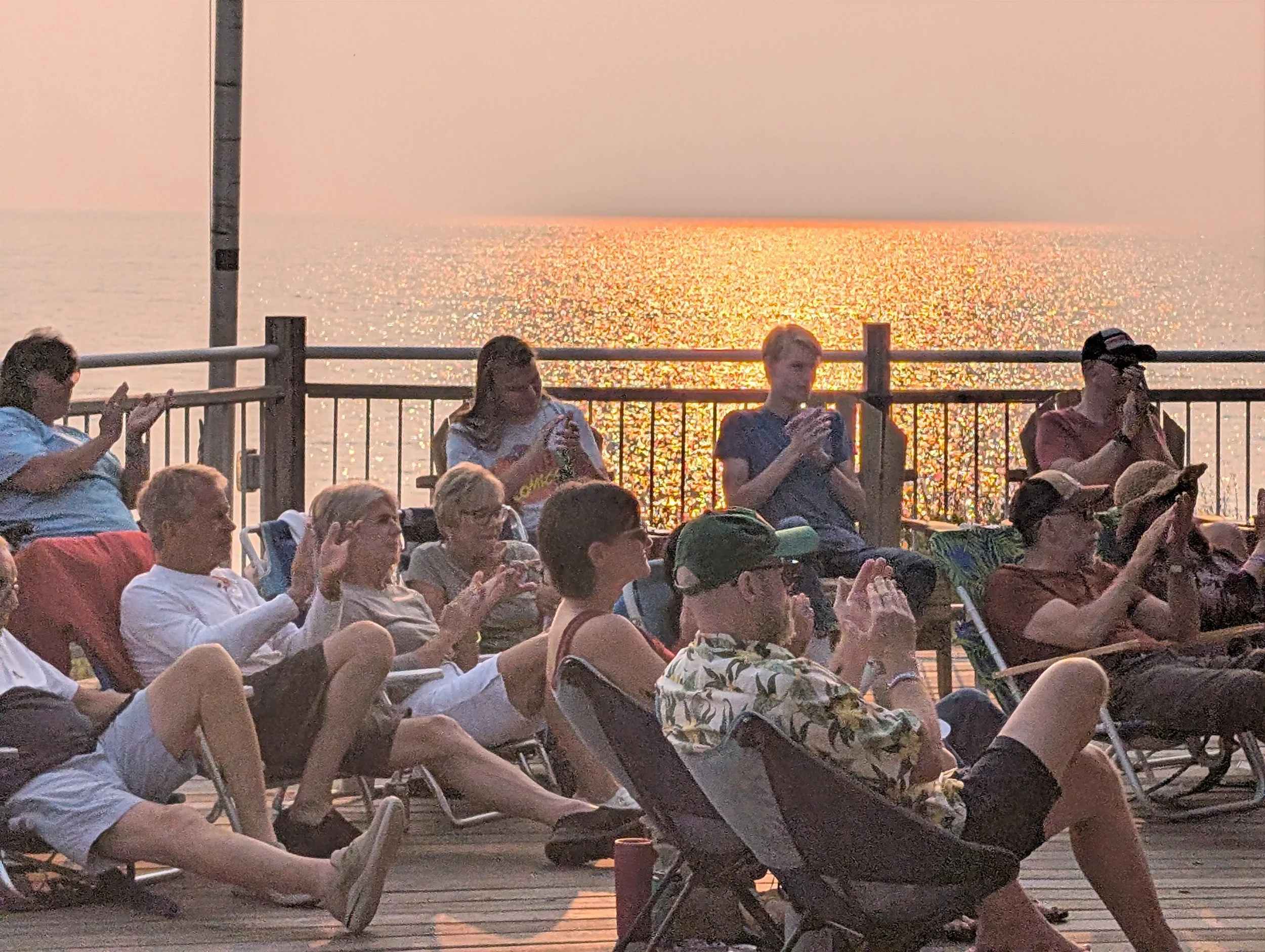 Group of people sitting on deck chairs and clapping, watching a sunset over the ocean.